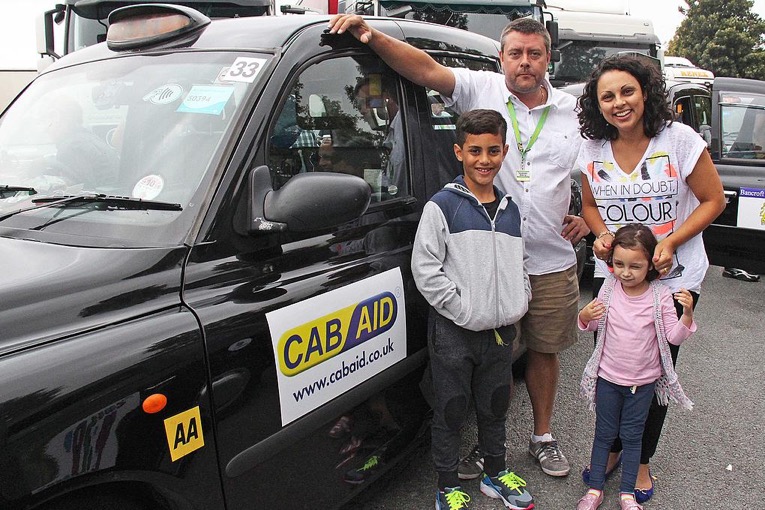 Two children and their mother stand aside a black cab smiling happily with the driver.