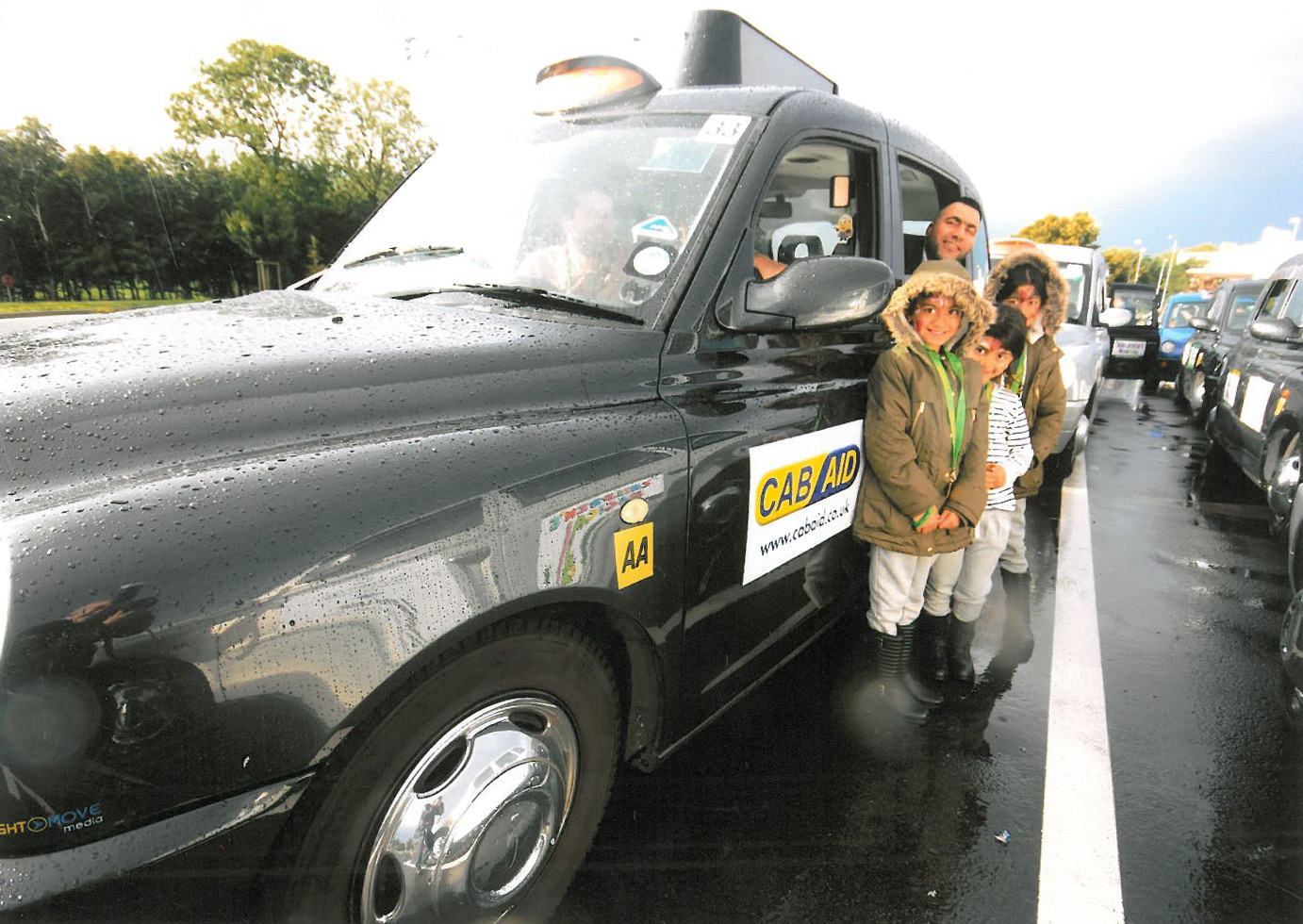 Three children stand, smiles brimming, in a sea of taxi cabs. A parent hangs out of the nearby car smiling for the camera.